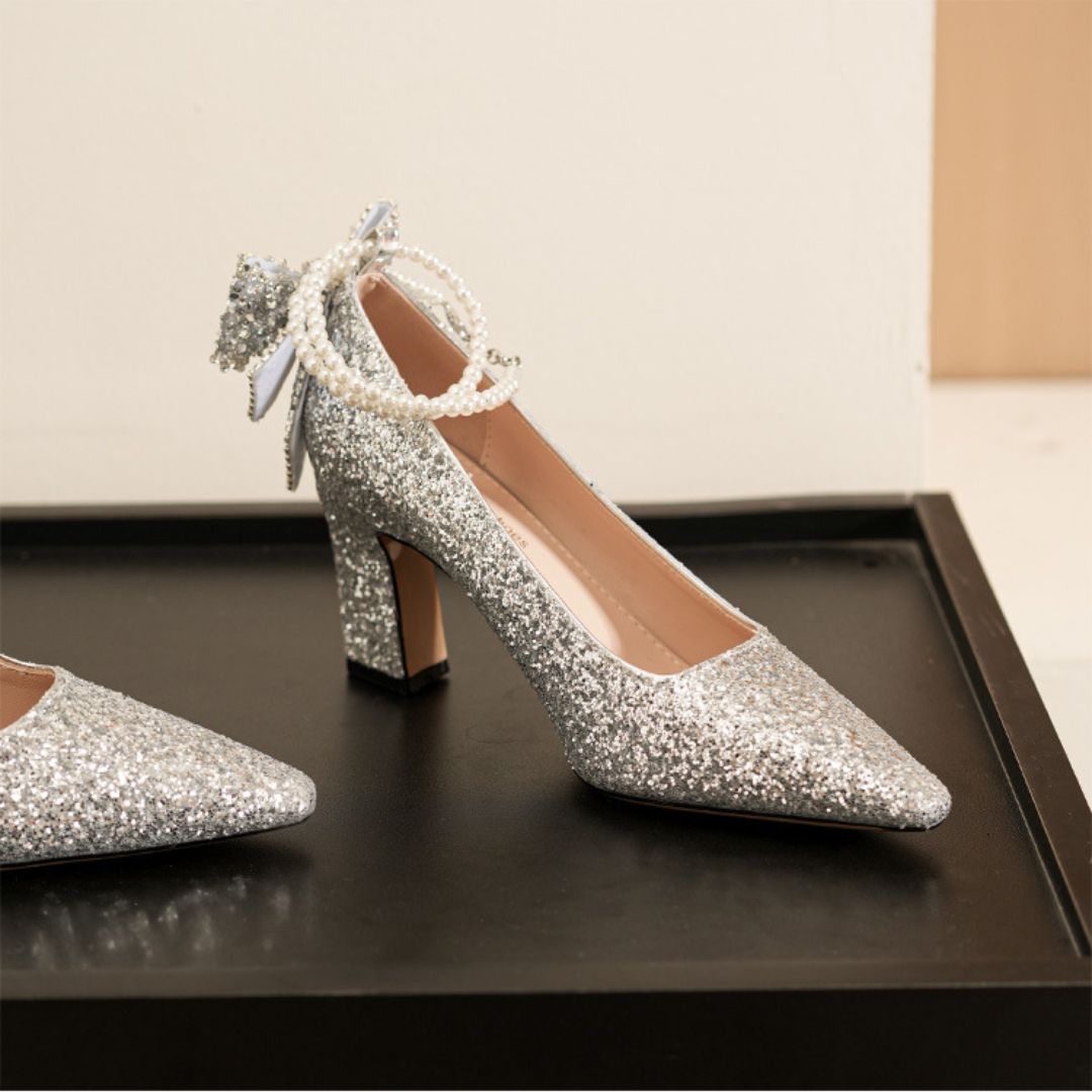 Silver glitter high-heeled shoes with pearl anklets on a dark surface.